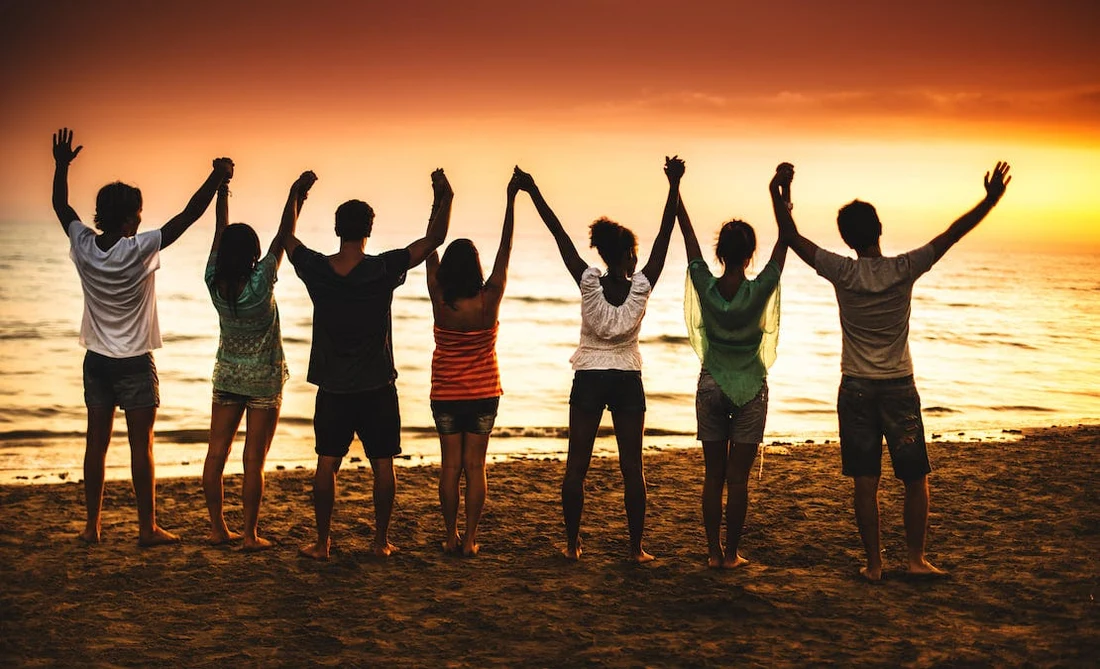 A group of friends holding hands, facing a sunset together
