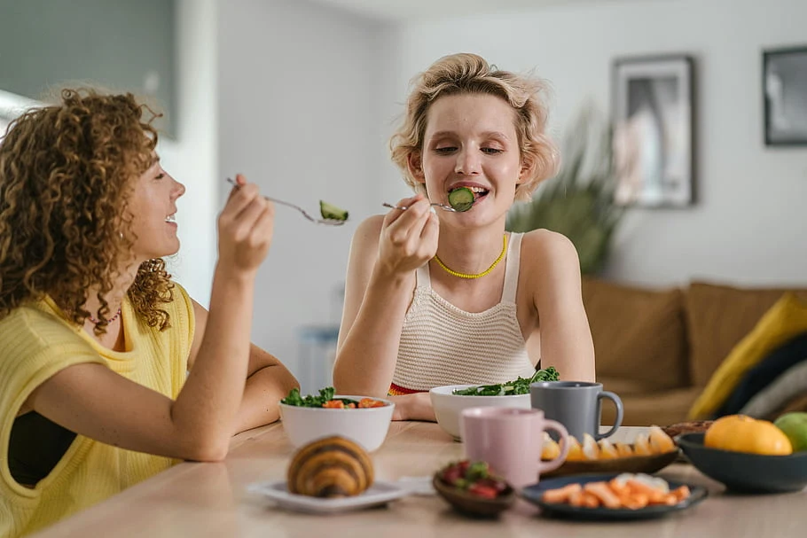 Two women laughing over a shared breakfast of greens, fruit, and eggs